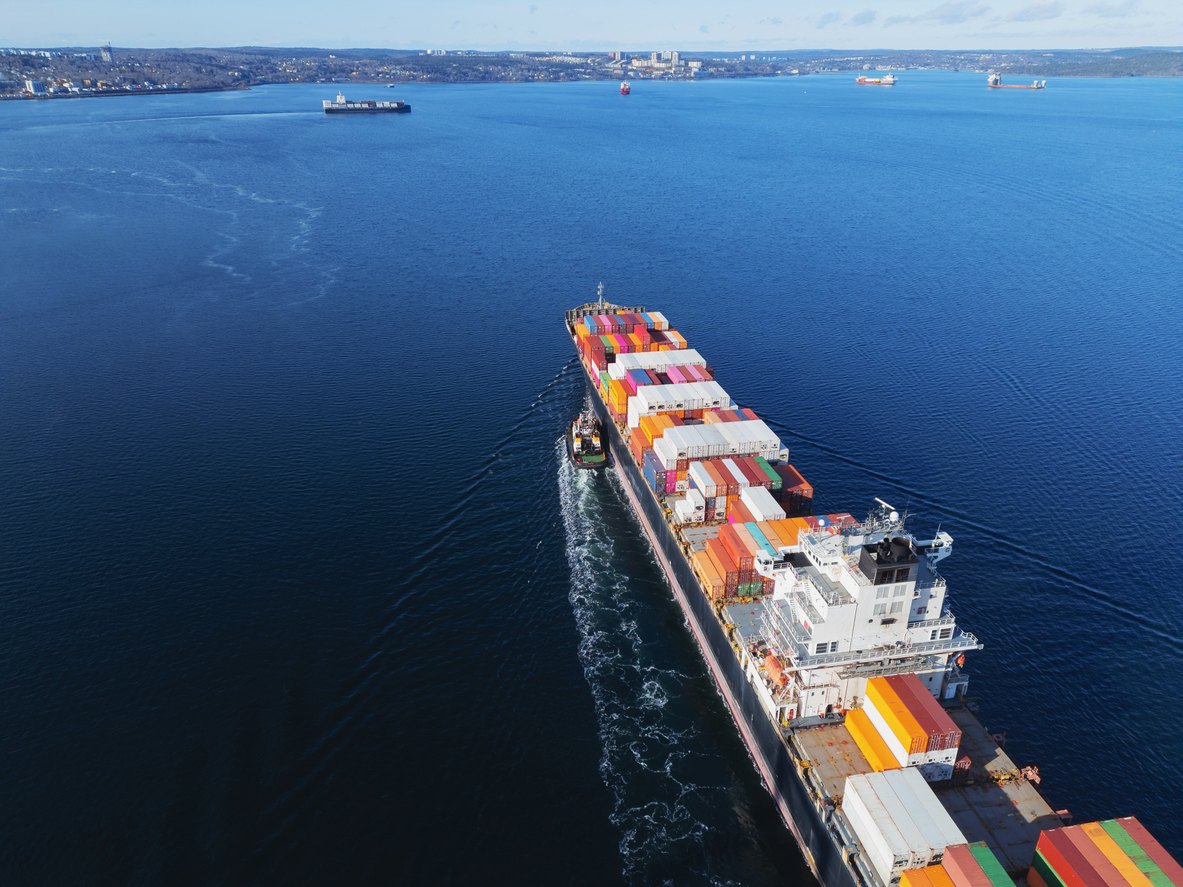 Aerial view of a container ship nearing port with the assistance of a tugboat.