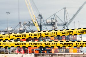 Yellow caution tape labeled "TARIFFS" in front of a shipping port with cargo containers and cranes, symbolizing trade restrictions.